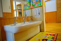 木の温もりを感じる室内に設けられた幼児用の三連水道の正面に大きな鏡、床には動物が描かれたマットが置かれ、右側の壁にはイラスト入りの手洗い手順が貼られている写真