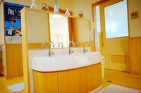 木のぬくもりを感じる室内に、子ども用の低い高さで設計された白と木目調の洗面台が並び、上には鏡と石けんが置かれている保育施設内の洗面コーナーの写真