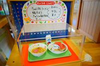 透明ケースの中にその日の給食がサンプルとして展示されており、ちゃんぽんラーメン、鶏そぼろ、すいか、牛乳がカラフルなトレイに盛られ、手書きのメニュー表が置かれている写真