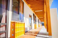 園舎の壁側に黄色の靴箱が部屋ごとに設置された、明るい太陽光が入る木の柱と屋根が印象的な園舎の外廊下の写真
