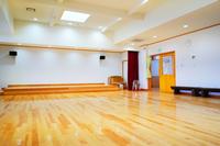 白い壁と明るい木の床が広がる、多目的に使えるステージ付きホールの写真