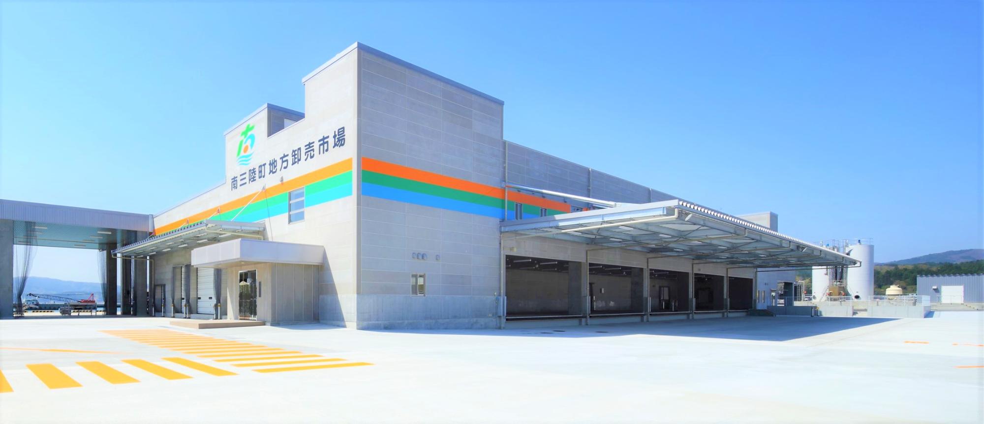 オレンジ、緑、水色のアクセントカラーが施されたグレー色の外壁に「南三陸町地方卸売市場」と書かれている市場で物流車両が待機・荷下ろしができるスペースや大きな庇が屋根から伸びている南三陸町地方卸売市場の外観写真