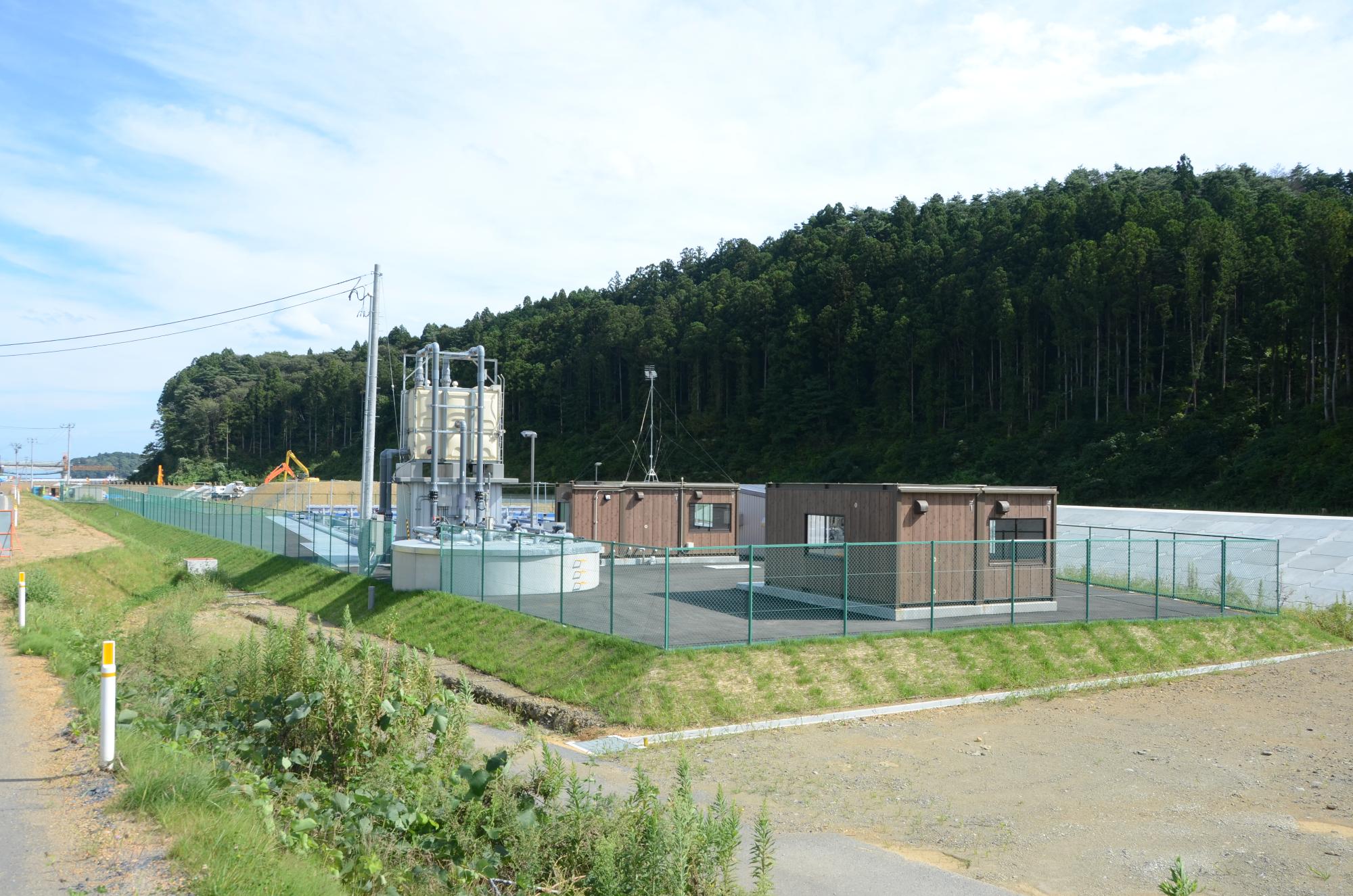 青空の下、山林の手前に緑色のフェンスで囲まれた敷地があり、複数の茶色い建物と白い円筒形の装置が設置された水尻ふ化場の外観写真