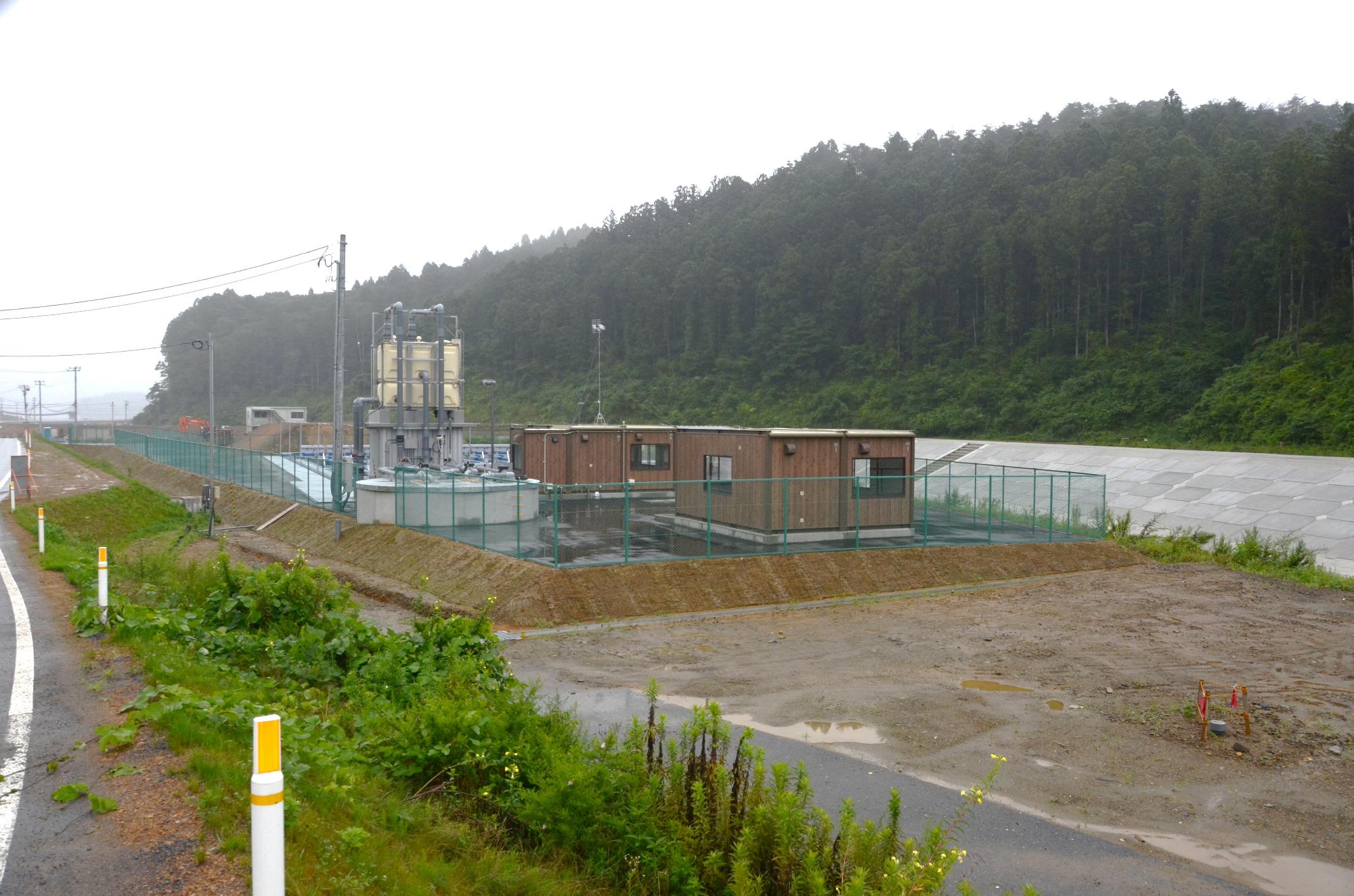 緑色フェンスで囲まれた敷地内に茶色い建物と白い大型装置が並んでいる完成間近の水尻ふ化場の写真