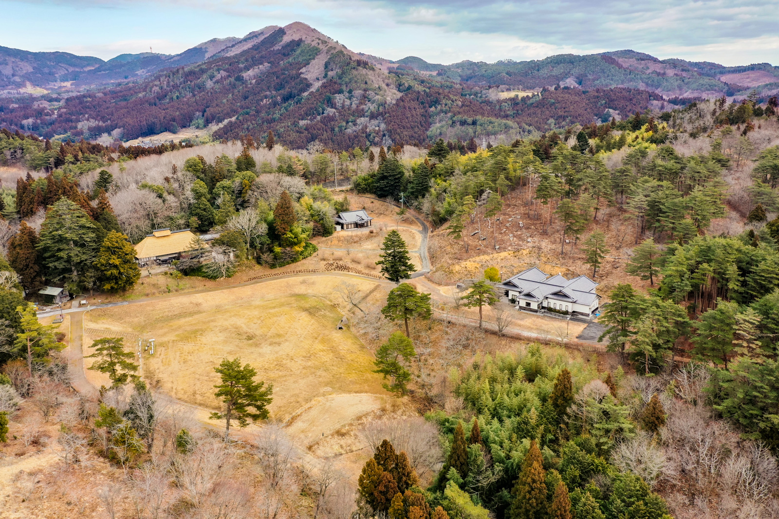 少し色づいた山々や木々に囲まれた広大な丘陵地帯の草地の中に、日本風の伝統的な建物と現代的な建物が点在し、その全景を上空から撮影した「ひうころの里」の風景の写真