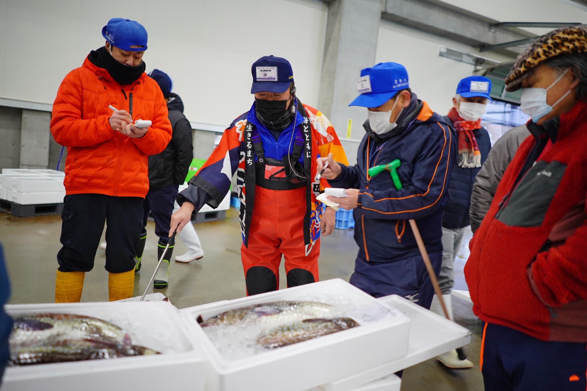 オレンジ色の法被を着た市場関係者が魚の入った発泡スチロール容器を指し示している様子の写真