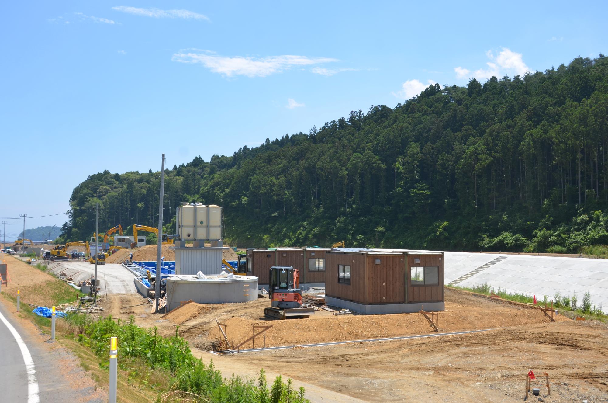 青空の下、造成地に設置された茶色い建物と白い大型タンクが並び、周囲では重機や作業員が作業する中、背後に緑豊かな杉林とコンクリート法面が広がる水尻ふ化場の建設中写真