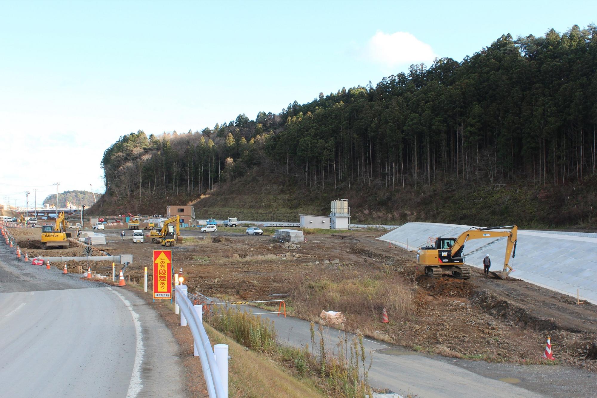 重機や作業員が稼働する広い造成現場で、道路沿いに工事用車両や重機が並んでいる水尻ふ化場の建設中写真