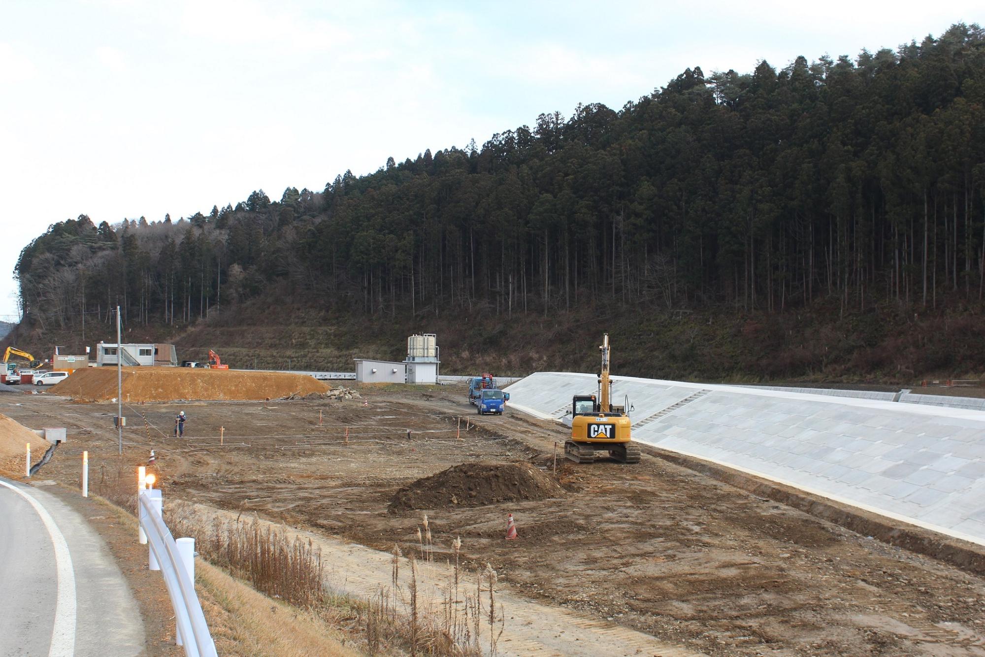 道路脇の造成地で黄色いショベルカーや青い作業車が稼働し、奥には仮設建物や白い設備がある水尻ふ化場の建設中写真