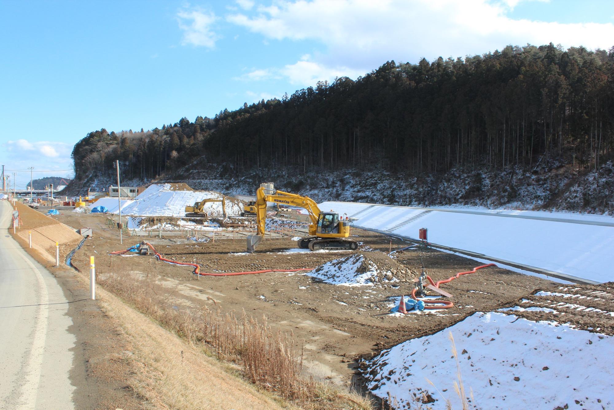 雪が部分的に積もる造成地で黄色いショベルカーが稼働し、赤いホースや資材が散在する水尻ふ化場の建設中写真