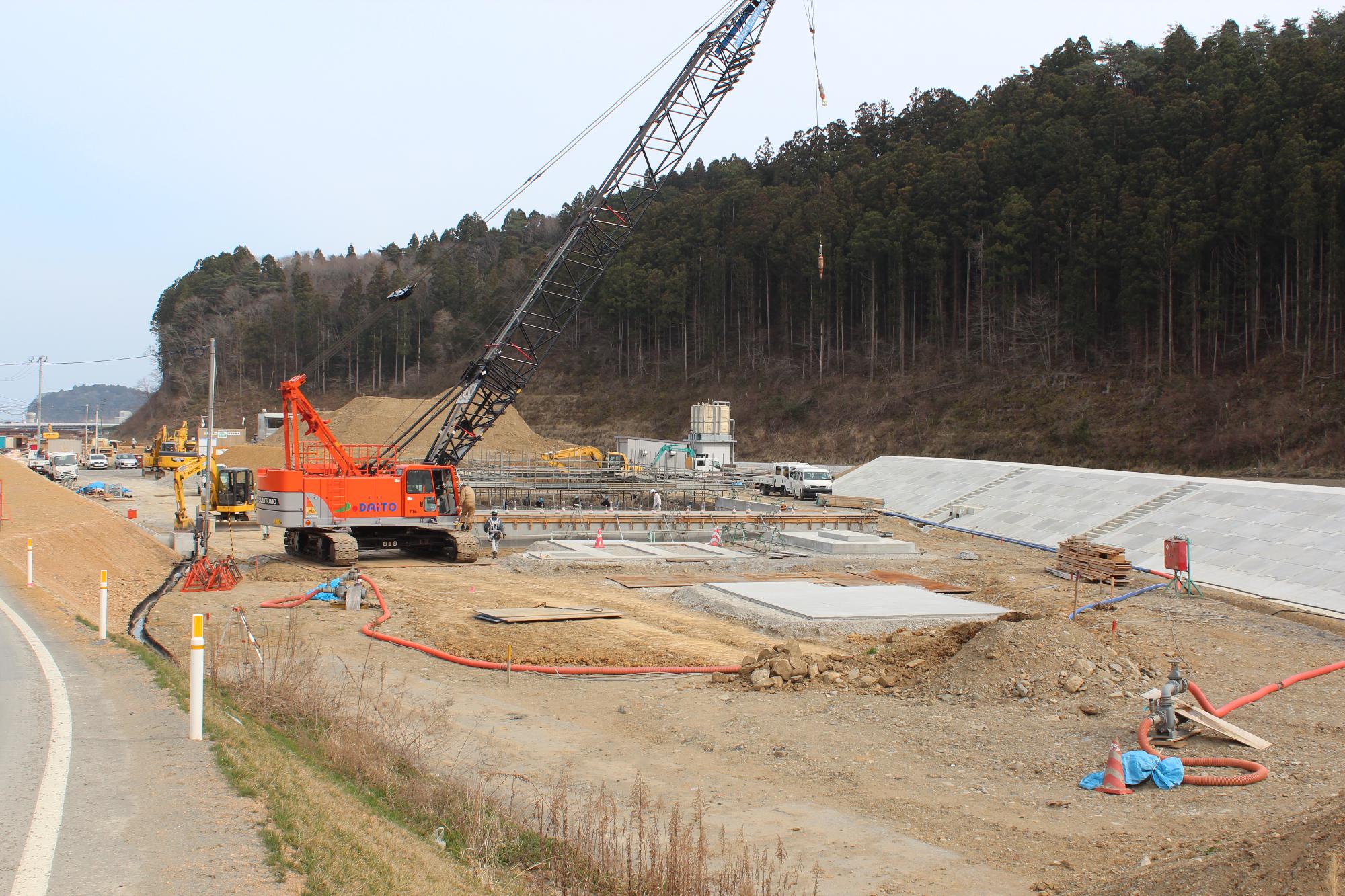 オレンジ色の大型クレーンが基礎工事中の造成地に配置され、コンクリートの基盤や資材が並んでいる水尻ふ化場の建設中写真