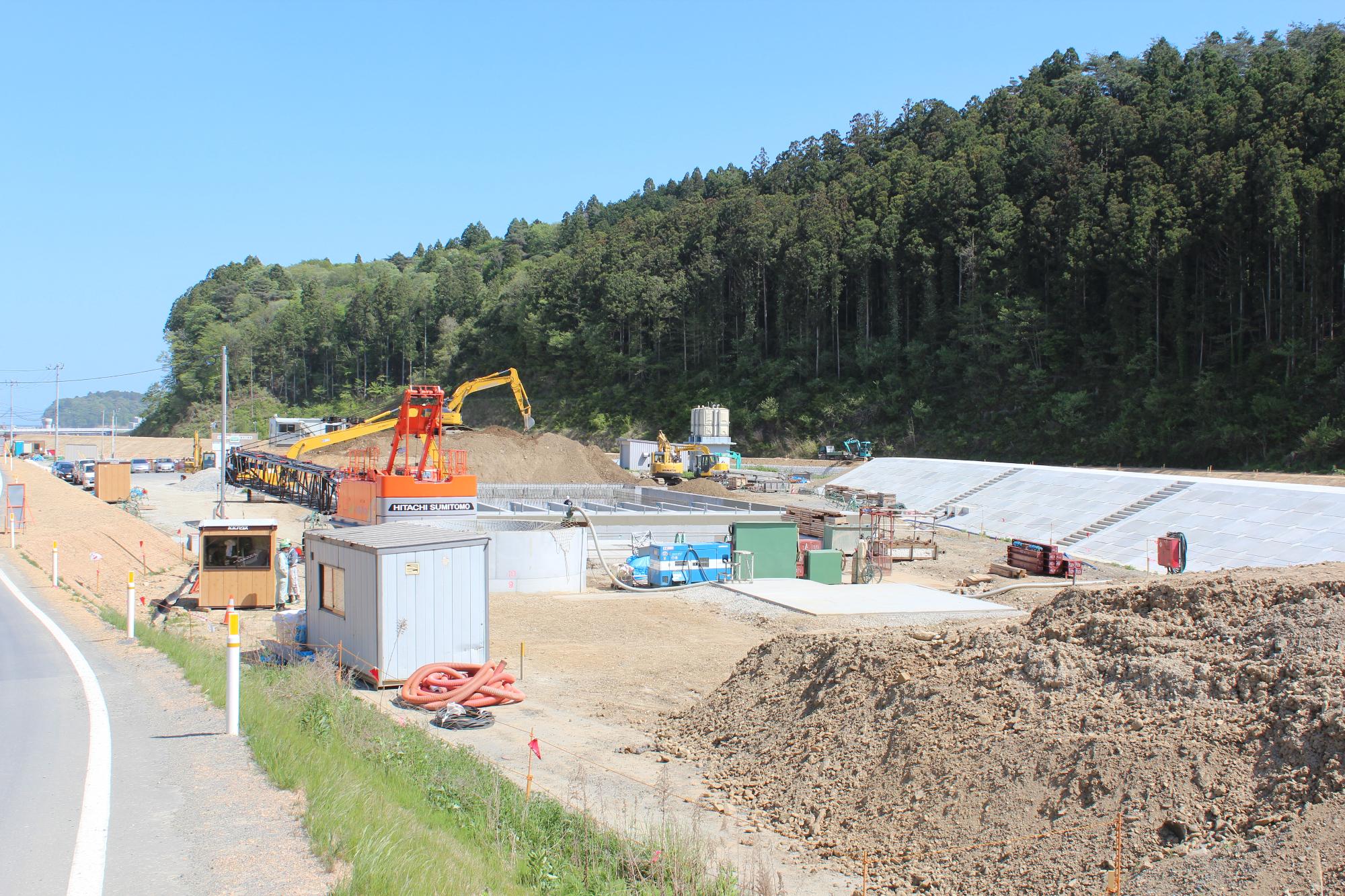 青空の下で大型クレーンやショベルカーが稼働し、資材や仮設小屋が並ぶ造成地にコンクリート基盤や盛土が見られ、背後には緑豊かな杉林が広がる水尻ふ化場の建設中写真