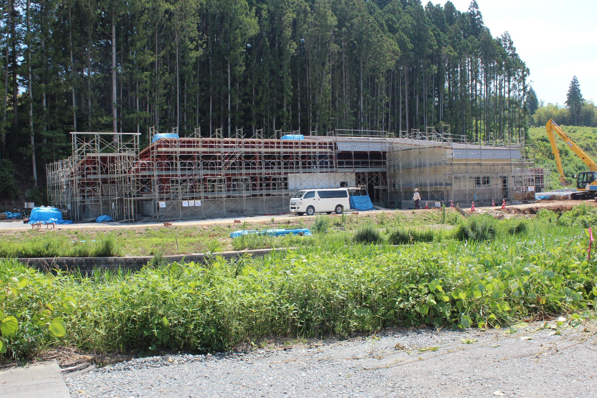 建設中の建物に足場が組まれ、白いワゴン車や作業員、右側には黄色いショベルカーが見える写真