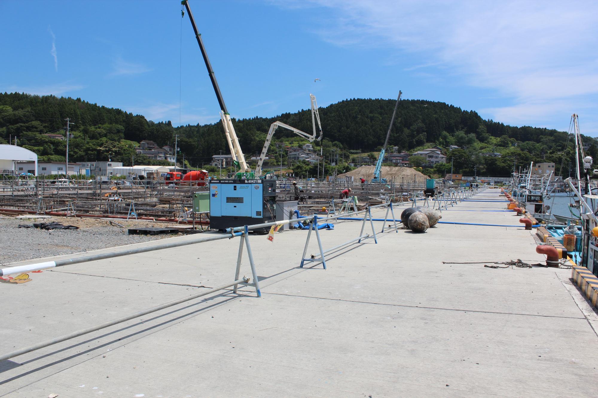 青空の下、港の岸壁沿いでクレーンやポンプ車が稼働し、鉄骨や資材が並ぶ中で建設工事が進む様子を捉えた写真