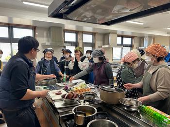 大きな調理台を囲んで複数の人々がエプロンと三角巾を着け、鍋や野菜を前に講師の説明を受けながら楽しそうに調理実習を行っている様子の写真