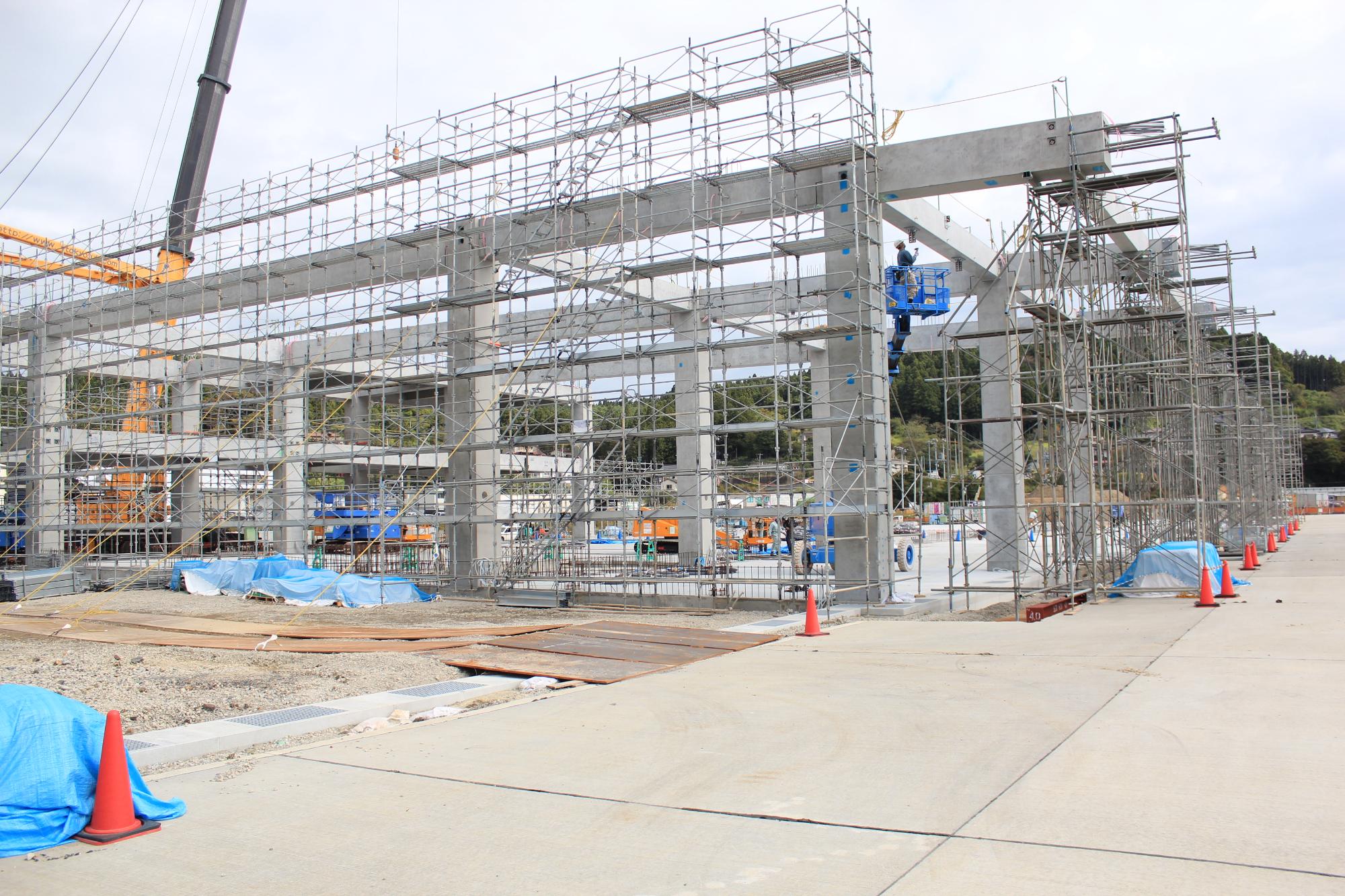 鉄骨と足場が組まれた建設中の建物の工事現場で、地面には青いシートとオレンジ色のコーンが配置されている写真