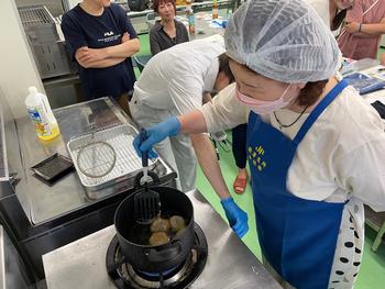 ヘアキャップを着けた女性が手袋をはめ、黒い鍋で揚げ物をひっくり返しながら調理実習を行っている様子の写真