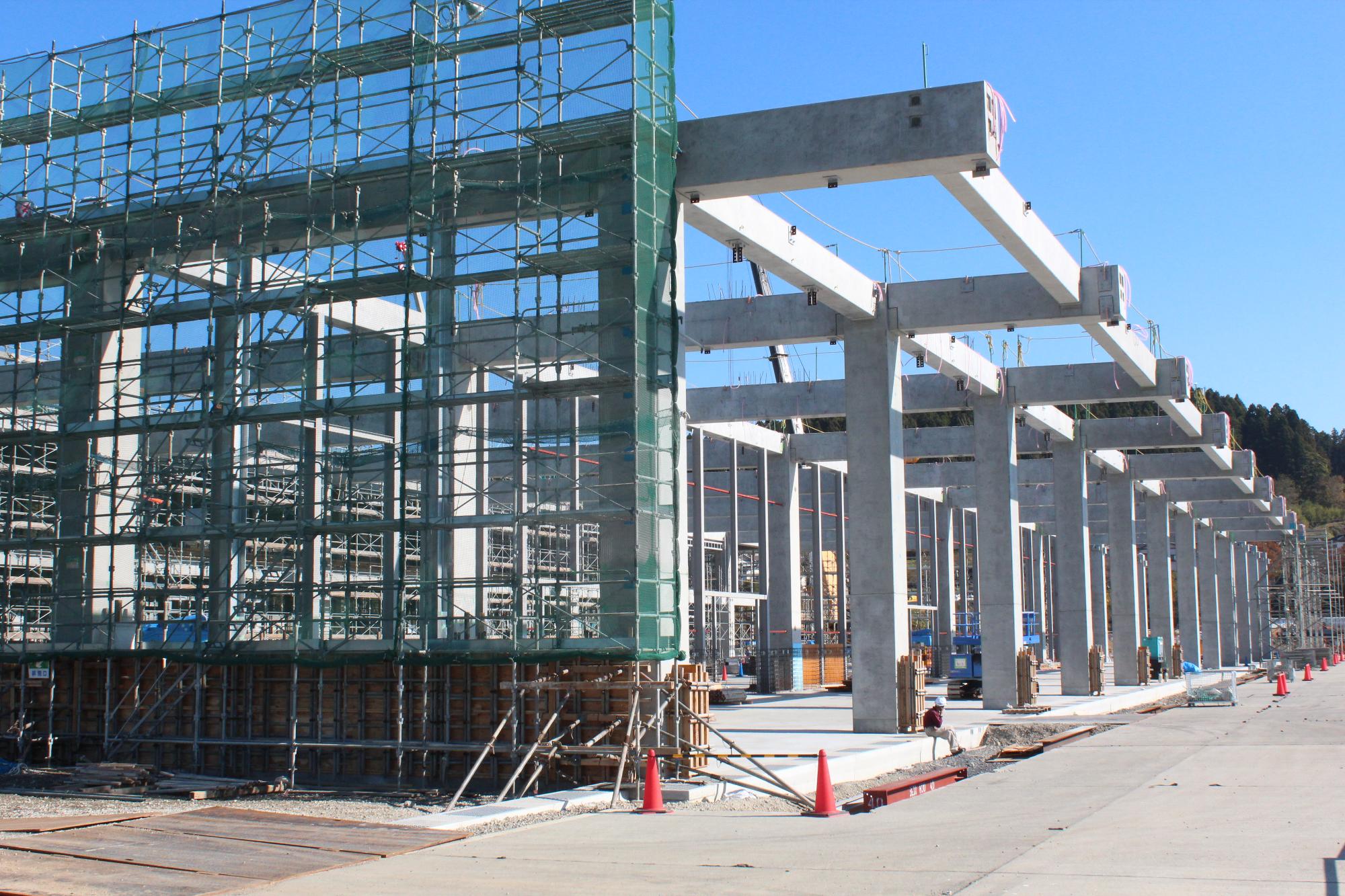 建設中の大きな建物の骨組みが見え、左側には足場、右側にはコンクリート製の柱と梁が並び、地面にはオレンジ色のコーンや資材が置かれている建設中の写真