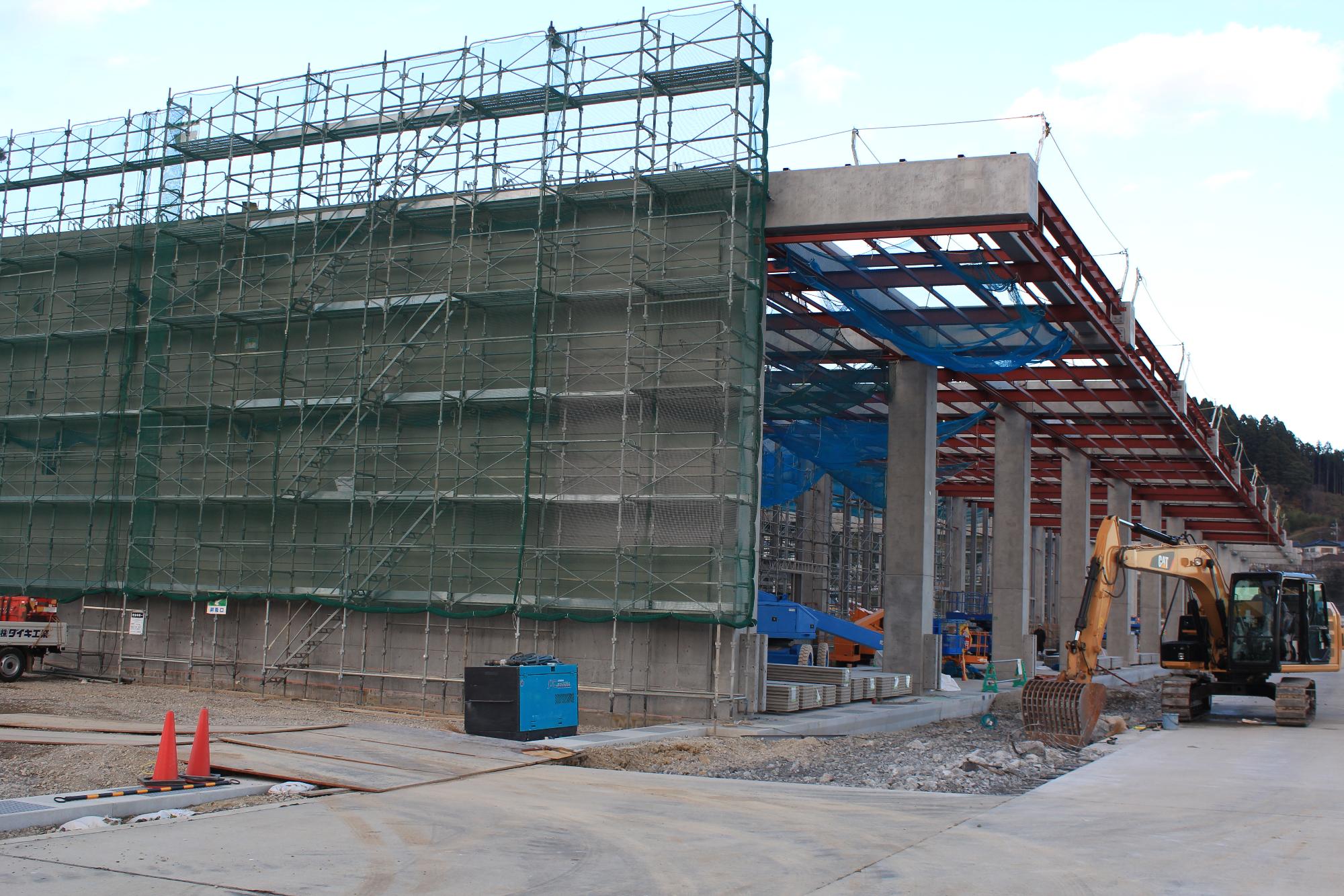 コンクリート柱が並び、上部には赤い鉄骨が組まれ、左側には足場と作業員用の青いネット、手前には工事用車両や重機が配置されている写真