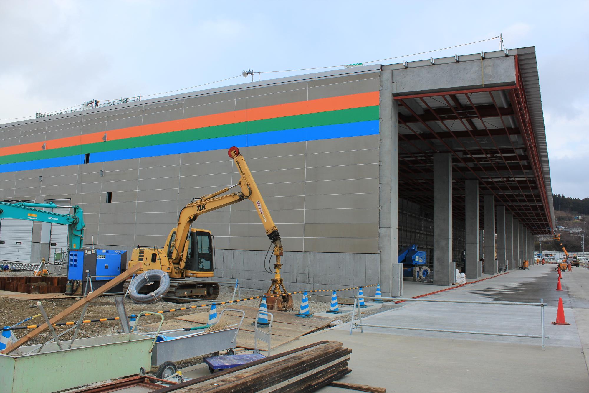 オレンジ・緑・青の3本線が描かれた壁が出来上がった建設中の市場の前に黄色い油圧ショベルが置かれている写真