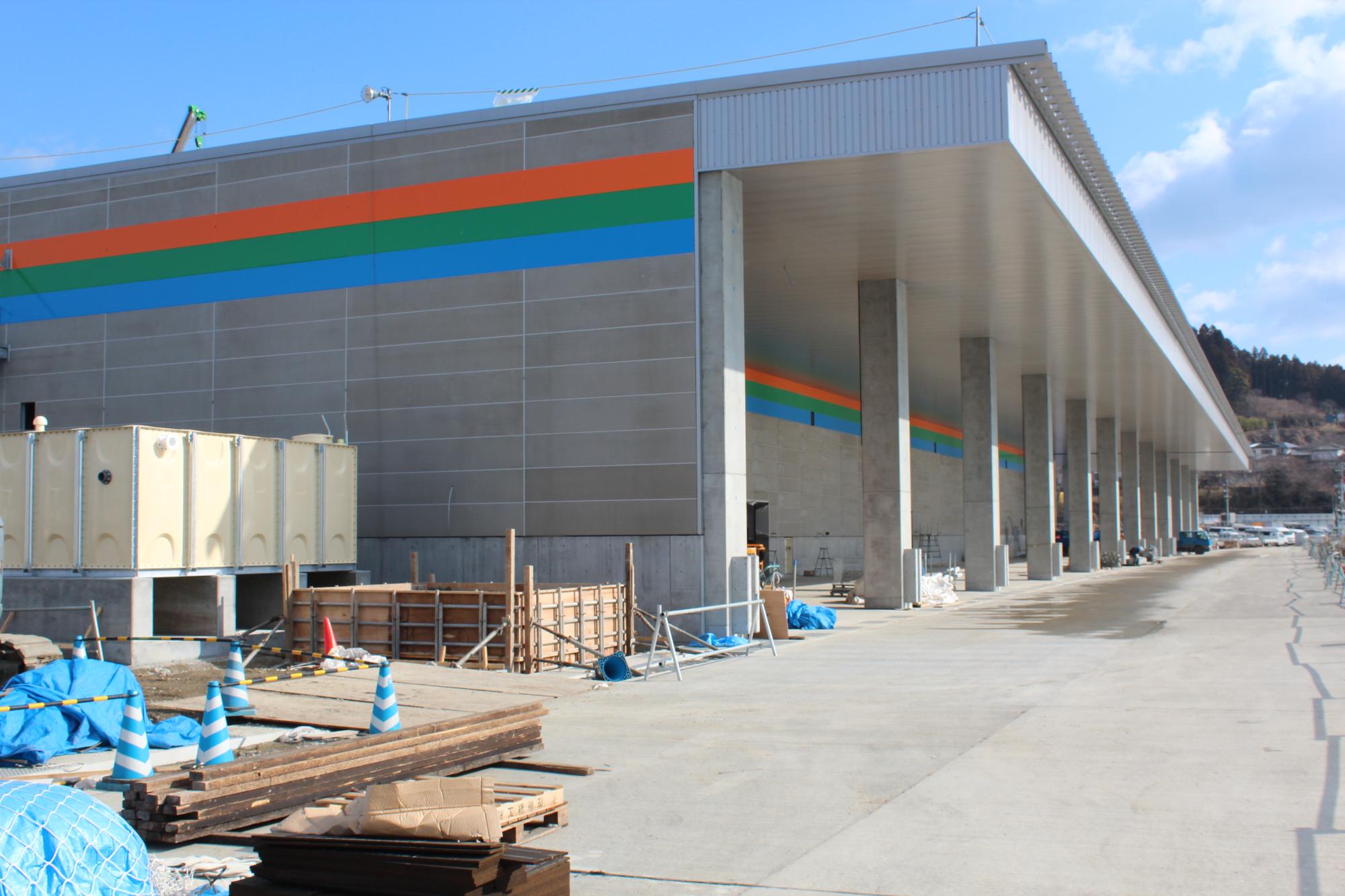壁にオレンジ、緑、青の3色の横線が描かれ、金属製の屋根がある市場の前に工事用資材や青いシートで覆われた物が置かれている写真