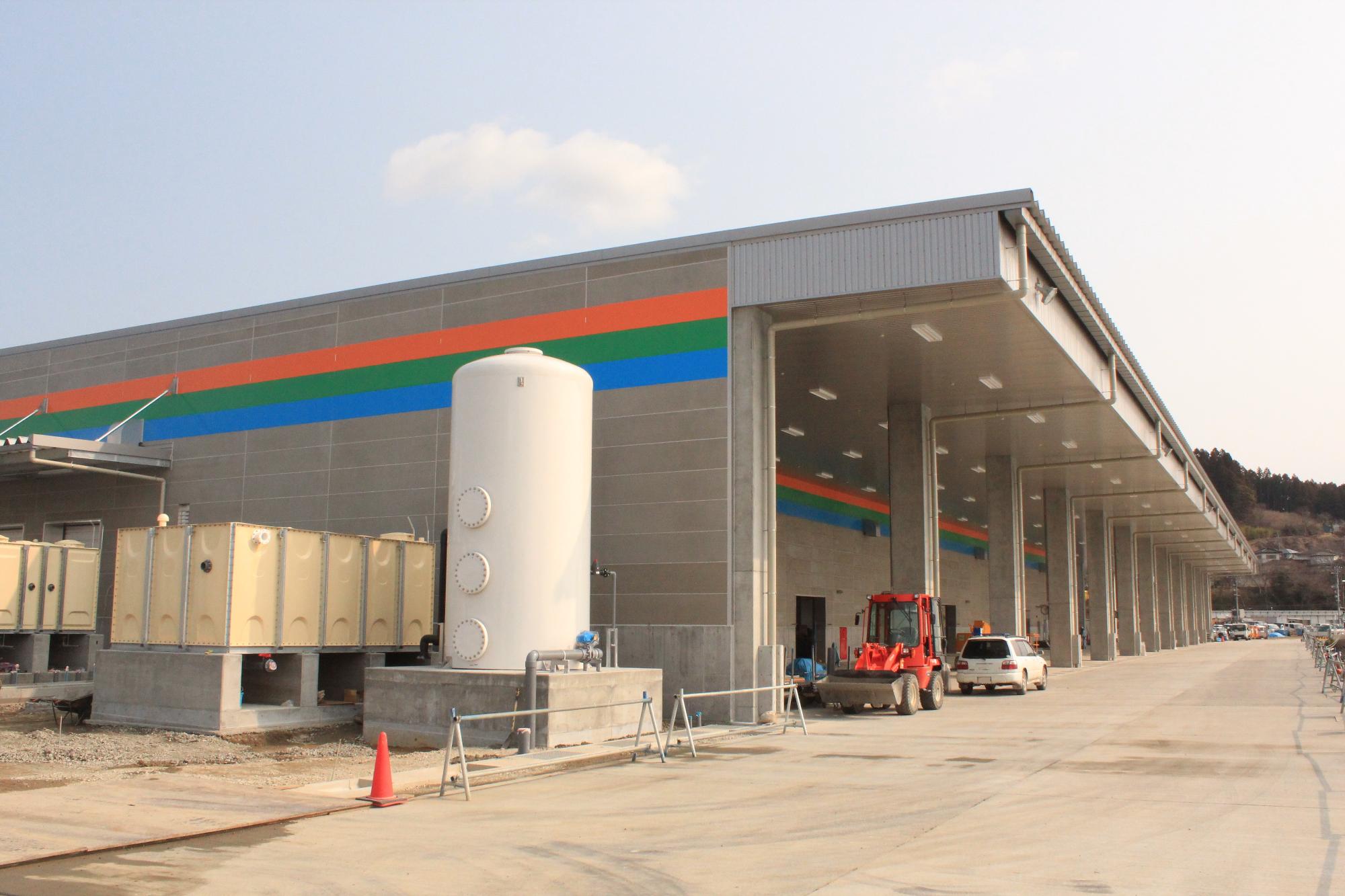 広い駐車場に面した大きな建物で、壁には緑、オレンジ、青の3本線が描かれており、白い円筒形のタンクや赤い重機が見える建設中の写真