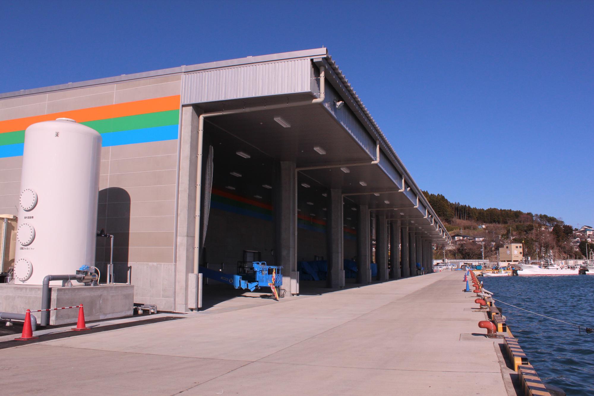 港の岸壁に沿って建てられた大きな建物で、外壁にはオレンジ、緑、青の3色のラインが描かれ、左側には大きな白いタンクがある完成間近の市場の写真