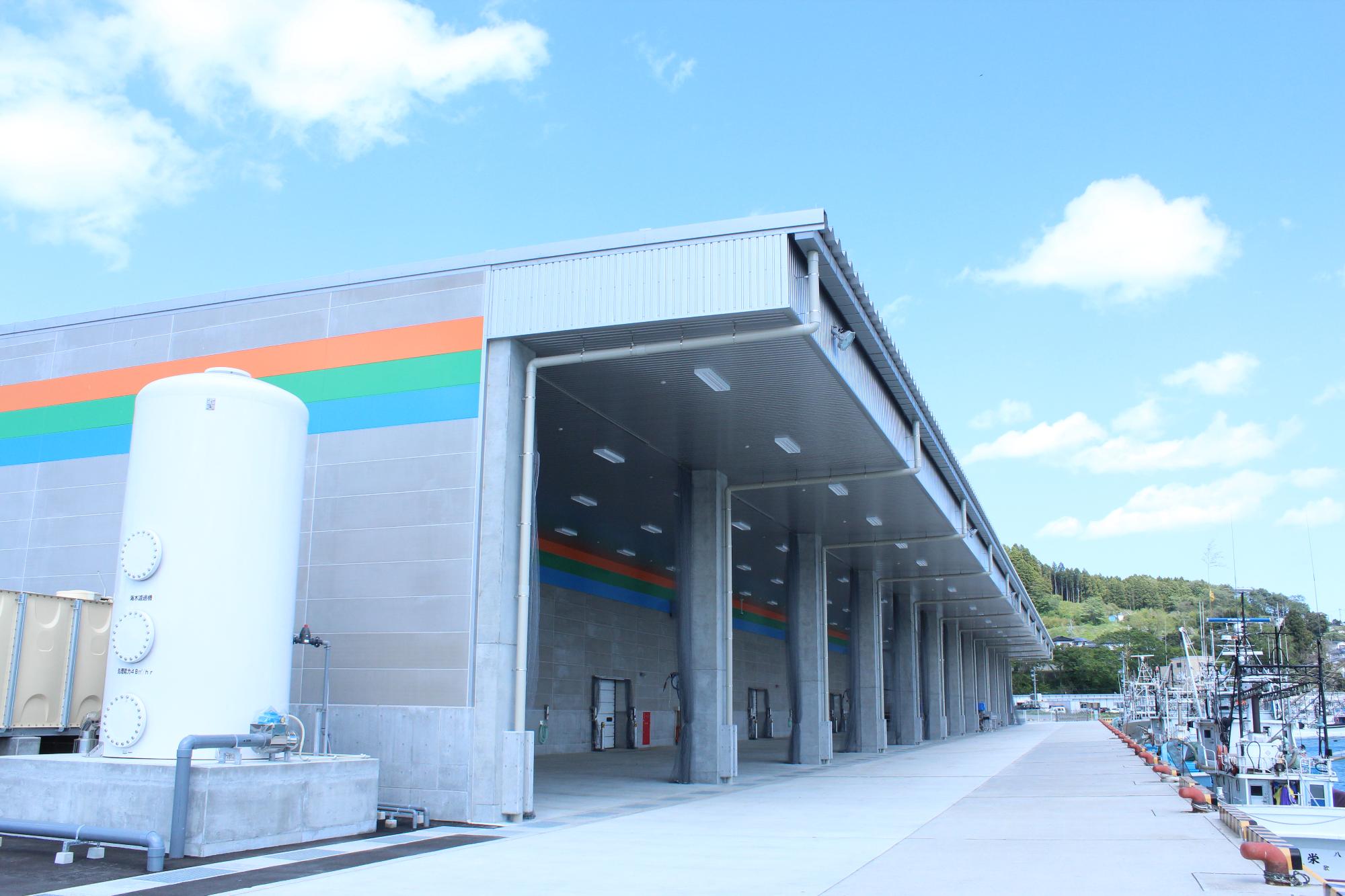 青空の下、壁にオレンジ・緑・青のラインが描かれた大型倉庫の横に白いタンクが設置され、右側には漁船が並ぶ港が続く南三陸町地方卸売市場の外観写真