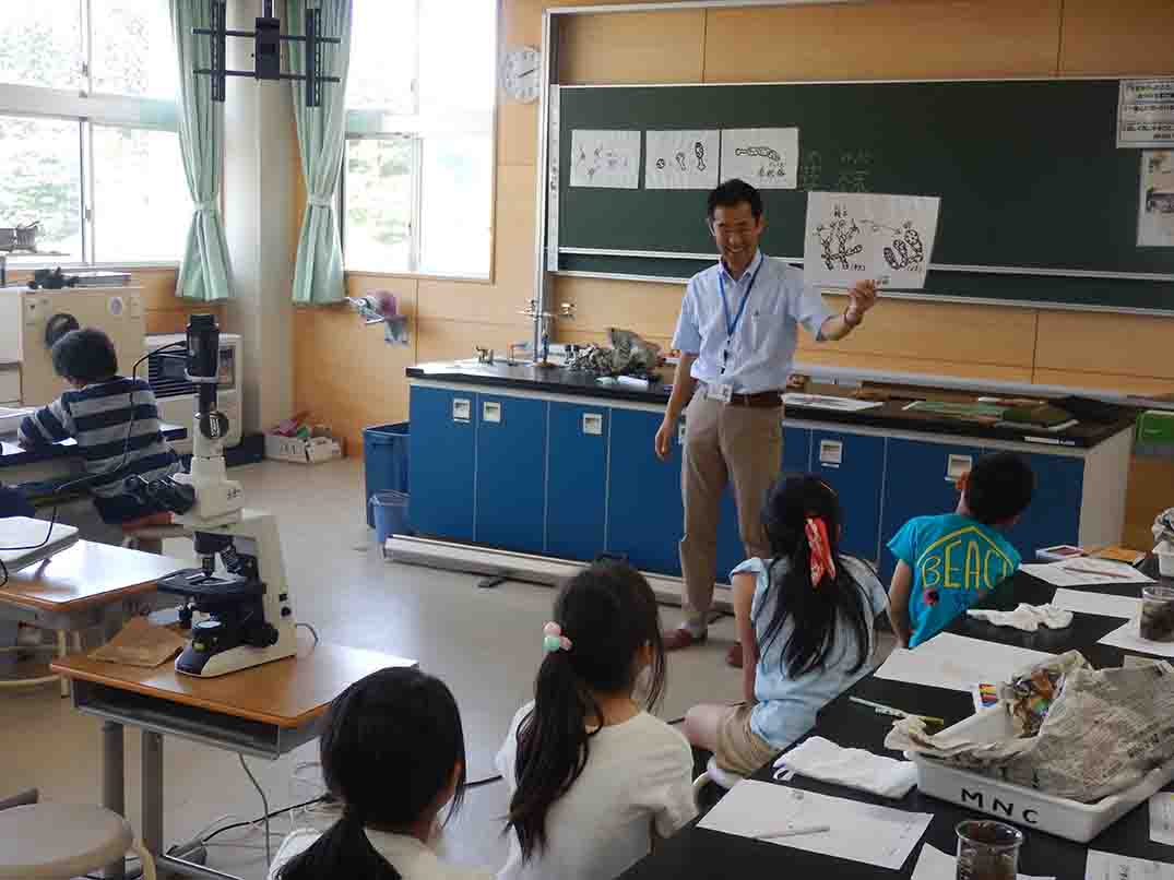 イラストが描かれた白紙を持った笑顔の男性が話しているのを、自席に座った子供たちが熱心に聞いている写真