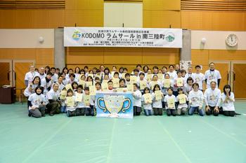 体育館に掲げられた「KODOMO ラムサール in 南三陸町」と書かれた白い横断幕の下で、大きなトロフィーの絵の前に整列し、修了証書を手に持った大勢の子どもたちと大人たちが並んで立っている集合写真