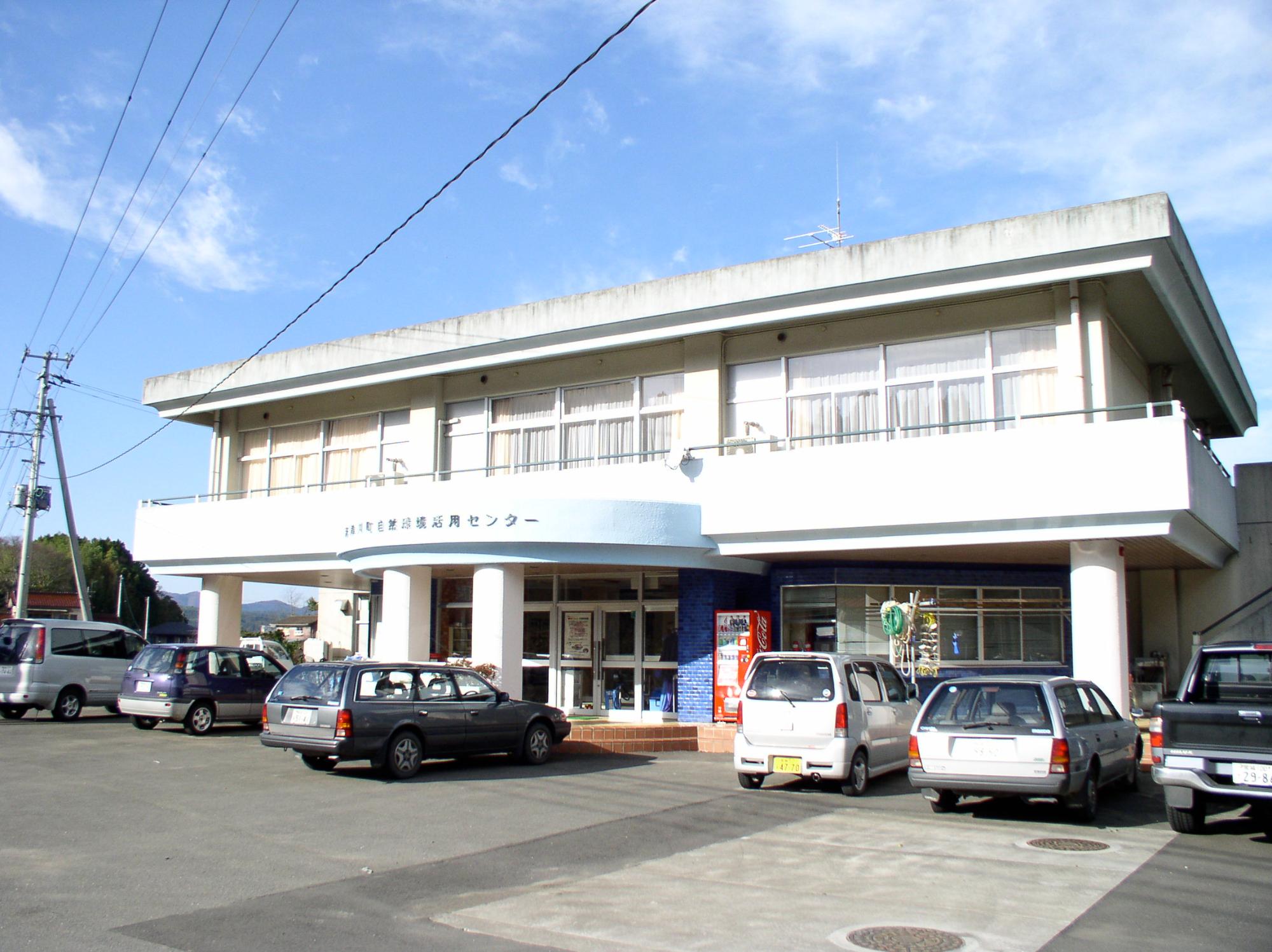 コンクリート製2階建ての建物が正面にあり、手前の駐車場には数台の乗用車が停まっている震災前の自然環境活用センターの外観写真