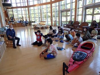 全面ガラス張りの屋内に設置されたカヤックの横に座ってガイドの男性の話を聞く子どもたちの写真