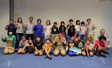 室内の青いカーペットの上で、前列中央に座ったココリコの田中直樹さんと一緒に、子どもたちや関係者が前後2列に並んで笑顔写った集合写真