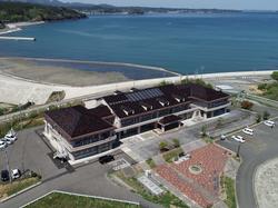 茶色い瓦屋根を持つ二階建ての大きな建物が手前にあり、その向こうには湾と防波堤、そして青い海が広がる航空写真