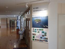室内の廊下の壁に、「自然環境活用センター」の案内板と上空から見た島の写真と複数の小さな風景写真と地図が並べられたポスターが張られている写真
