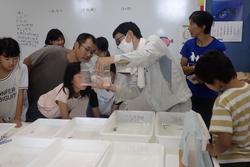 室内で生き物が入った袋を見せている大人と子どもたちの写真