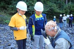 ヘルメットをかぶった子どもたちが川原で講師の話を聞いている写真