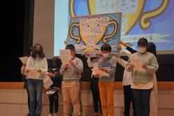 後ろに大きなトロフィーの絵が掲げられているステージ下で、紙を持った子供たちが発表の内容を読み上げている写真