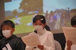 白い服の女の子がマイクを使って真剣に発表する女の子の写真