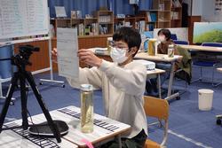 教室でカメラの前に座った子どもが、資料を掲げながらオンライン発表の準備をしている写真