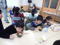 室内で子どもたちが机を囲み、味噌汁やご飯などの郷土料理を楽しそうに食べている写真