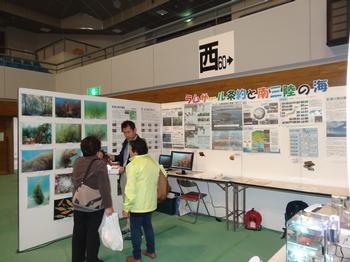 屋内施設で、ラムサール条約に関するパネルや写真が展示されたブースに、数人の来場者が集まり、タクゾー博士から説明を受けている様子を捉えた写真