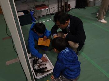 室内展示ブースの床で、白い容器に入った海藻を囲み、タクゾー博士と2人の子どもが熱心に観察している様子を捉えた写真