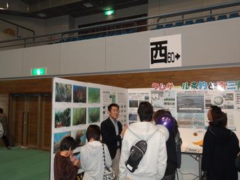 屋内で行われているイベント会場で、ラムサール条約に関するパネルの前で、数人の来場者がタクゾー博士と話しながら展示を見ている様子を捉えた写真