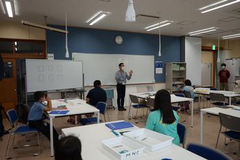 教室内のホワイトボードの前に立つ男性の話を子供たちが熱心に聞いている写真