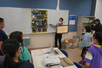 表彰状を持ちホワイトボードに貼られたポスターの前に立つ男性の話を、子供たちが立って聞いている写真