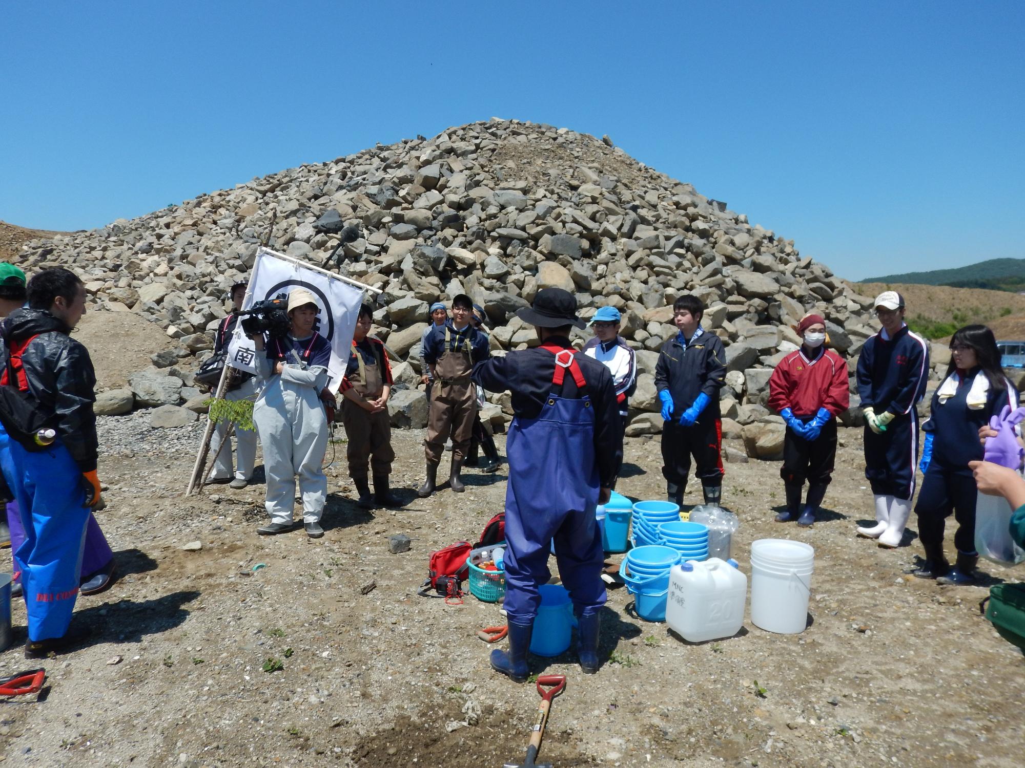 石が山積みされた屋外にバケツなどの道具が置かれた傍に立つ男性の話を、輪になった参加者が聞いている写真