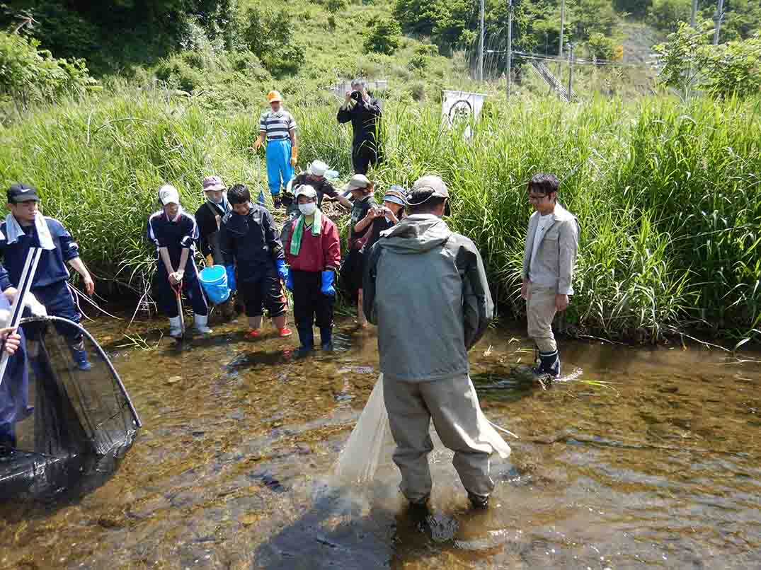 参加者たちが見ているなか、川の中に入った男性が水の中に網を沈めている様子の写真