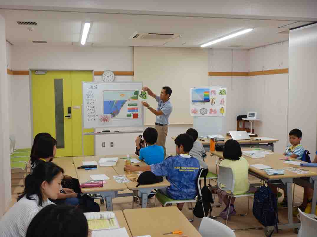 8人の参加者たちが室内前方で地図やイラストを使って説明している男性の話を聞いている様子を撮影した写真