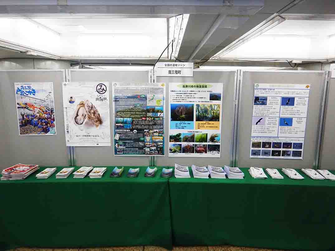 緑色の布がかけられた長いテーブルの上に、複数のパンフレットや冊子がきれいに並べられ、その後ろの壁には複数のポスターが掲示されている展示スペースの写真